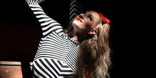 Cirqulinair 2026 - Deepfake Een acrobate in zwart wit gestreept pakje steekt haar been langs haar oor omhoog.