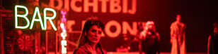Scenebeeld met Moeder Courage op de voorgrond, ze kijkt triest. Achtergrond in rood licht, de kar met de letters ‘Bar’