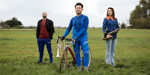 3 mensen in een weiland: een man in trainingsbroek met zijn handen in zijn zak, een man met een racefiets en bugeltrompet en een vrouw in een fanfareuniform met een saxofoon.