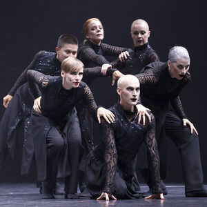 Zes dansers staan in een kluwen bij elkaar. Zij zijn in het zwart gekleed met kant en glimmers.
