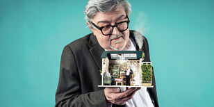 Acteur Beppe Costa heeft in zijn hand een klein poppenhuis, met een rokende schoorsteen. In het huisje staat hijzelf als poppetje. Ze kijken elkaar aan.
