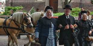 Twee in Westfriese kledingdracht gehulde dames lopen gearmd met een man met bolhoed uit voor twee paarden die een koets trekken.