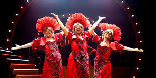 Drie oudere mannen gekleed in lange felrode galajurken en met veren hoofdtooien staan stralend in een showpose.