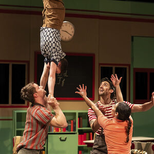Een acrobaat maakt een handstand op de handen van een andere acrobaat. Twee collega's staan klaar om hem op te vangen.