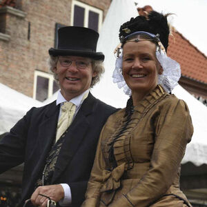 Op de bok van een open koets zitten een man met hoge hoed en een vrouw in Westfriese kledingdracht.
