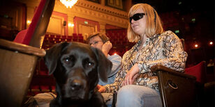 Een vrouw met een koptelefoon en een donkere bril zit in een theaterzaal. Op de voorgrond de kop van een hond.