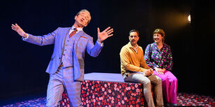 Paul Groot als de weddingplanner staat uitbundig te zingen. Achter hem zitten Bob en Jenny naar hem te kijken. 