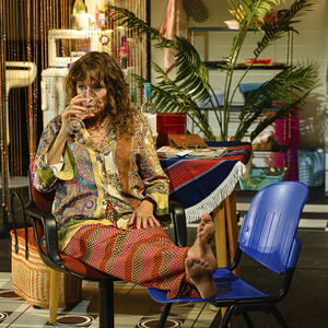 Renée Fokker neemt een slok wijn, zittend op een stoel in een gezellige huiskamer met veel planten. Ze heeft haar benen op een andere stoel gelegd.