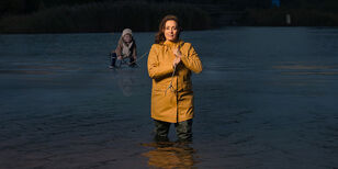 Een vrouw staat tot haar knieën in het water, een sleetje met kind voorttrekkend over ijs