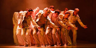 Een groep dansers strekt vooroverbuigend de hand uit naar rechts, in koperbruin licht.