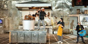 In een enorme chroomkleurige keuken staat een kok naast een mega kookpot met een orang oetan erin