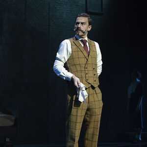  Hercule Poirot  in hemdsmouwen, tweed vest en -broek kijkt met een gefronst voorhoofd opzij. In zijn hand houdt hij met een wit zakdoekje een mes vast.