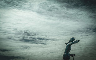 beeld van wijzend oud vrouwtje met een stok, voor een grauwgrijze wolkenlucht