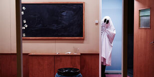 Een als spook verkleed persoon kijkt om het hoekje van een klaslokaal. 