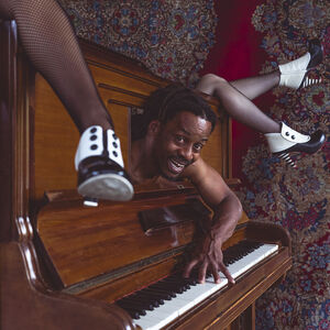 Het lachende hoofd van een man met dreadlocks steekt uit de voorkant van een piano, uit de bovenkant van de piano steken drie vrouwenbenen.