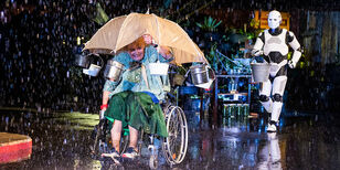 Betty in rolstoel onder paraplu waar metalen emmertjes aan hangen, op de achtergrond komt robot Shiny met een grote emmer aanlopen