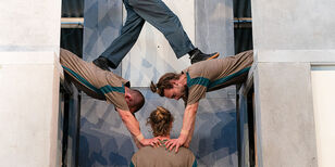 Twee acrobaten leunen links en rechts over een muur. Met hun handen houden ze de schouders van een derde acrobaat vast. Een vierde acrobaat staat op de ruggen van de leunende acrobaten