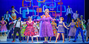 Het grote ensemble van Hairspray, dansend in vrolijke jaren 60 kleding. Middenvoor staat Richard Groenendijk als moeder Edna in een enorme paarse jurk.