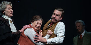Een huilende vrouw met een rozenkrans in haar hand, duwt beide vuisten tegen de borst van Poirot. Een net geklede oudere vrouw legt haar hand op de schouder van de huilende vrouw.
