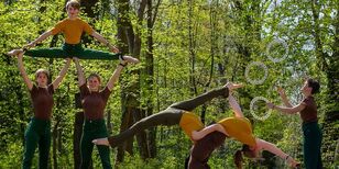 Acrobatische tieners in bruingroene kleren in het bos. Een meisje gooit 3
ringen in de lucht en twee meisjes tillen iemand die spagaat doet boven hun
hoofd.