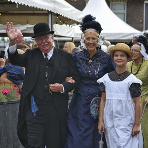 Gekleed in Westfriese kledingdracht lopen een oudere man en vrouw gearmd over een markt, samen met een meisje in een wit schort.