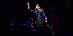 Sjors van der Panne als Jacques Brel staat achter een microfoon en zingt. Hij houdt daarbij een arm omhoog. 