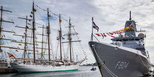 Een groot zeilschip met vier hoge masten. Ernaast een grijs marineschip, versierd met vlaggetjes.