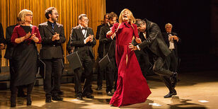 zangeres in rode lange avondjapon wordt lachend het toneel opgeduwd terwijl het koor klappend toekijkt