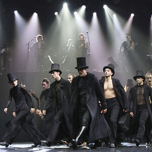 Lopende groep dansers met lange zwarte jassen en hoge hoeden. Door de lichtstralen in de verte staat de band Blaudzun.
