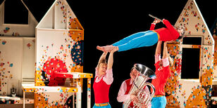 Een man en vrouw houden een vrouw boven hun hoofd vast. Zij speelt de dwarsfluit, terwijl onder haar een man tuba speelt. 