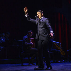 Sjors van der Panne als Jacques Brel staat achter een microfoon en zingt. Hij houdt daarbij een arm omhoog.