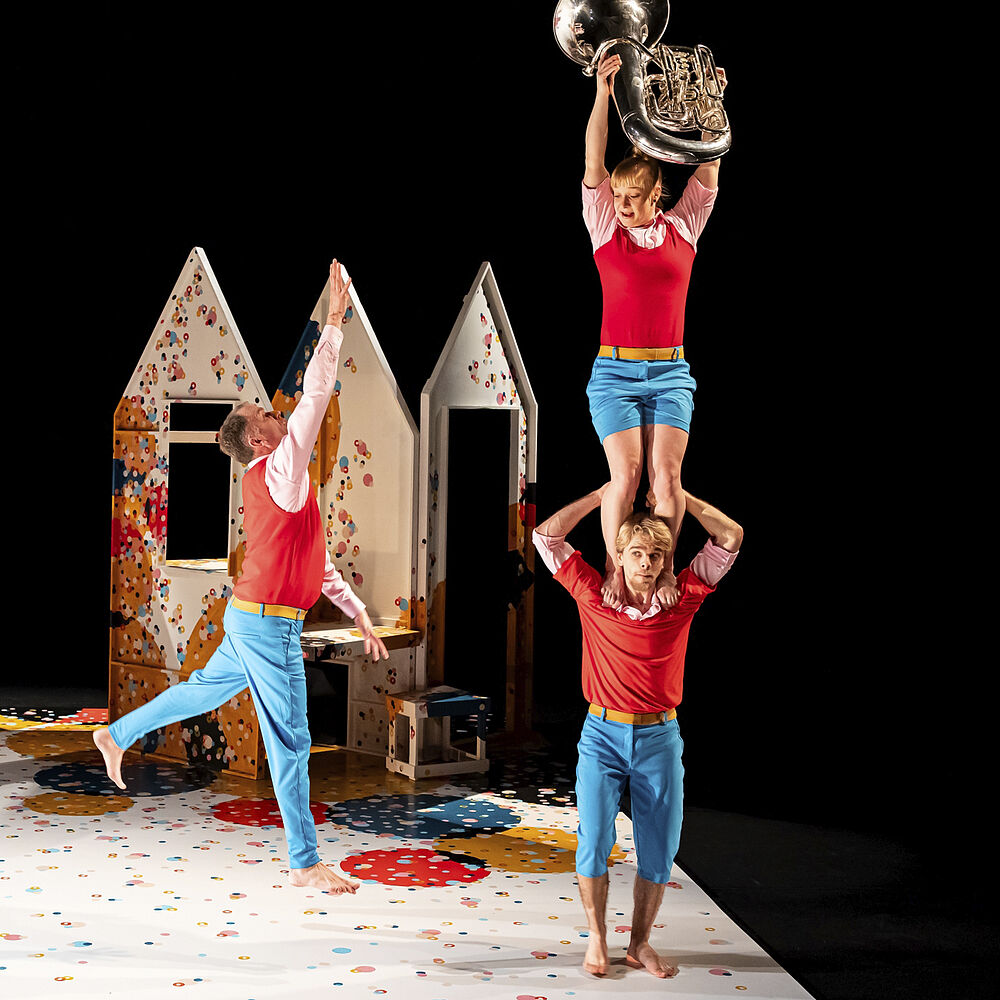 Een acrobate staat op de schouders van een man en houdt een tuba hoog boven haar hoofd. Een man springt om erbij te kunnen.