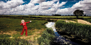 Karin staat zingend in een weiland. Ze draagt een rood pak.