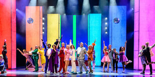 20 spelers in showposes op het toneel. Op de achtergrond een decor met brede strepen in regenboogkleuren.
