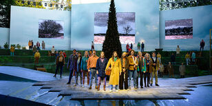 Alle acteurs staan in een v-vorm op het grote podium te zingen. Op de achtergrond een verlichte kerstboom.