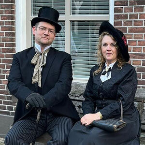 Een man en een vrouw zitten op een bankje voor een huis. Zij dragen zwarte kleding uit de 19e eeuw en hebben allebei een hoed.
