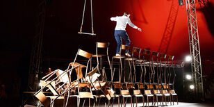 acrobaat loopt al balancerend over een rij wankel opgestapelde stoelen