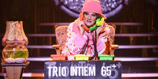 Mr Eddy 06 in roze peignoir en muts zit in bruine leunstoel achter tafeltje met telefoons en de tekst Trio Intiem 65+