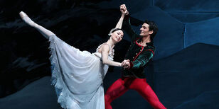 Een ballerina in een witte jurk steekt haar been omhoog en wordt hierbij ondersteund door een danser in een rood en groen kostuum. 