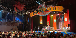 Mensen in traditionele boerenkleding staan op het podium Boven het podium hangt de slogan 'Agge mar leut et'. Het publiek is verkleed in boerenkleding, gehaakte vitrage en hebben lampenkappen als hoofddeksel. 