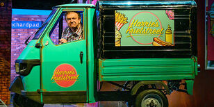 Jon van Eerd zit als Harrie in een groen tuktuk autootje met daarop reclame van Harries Frietstreet