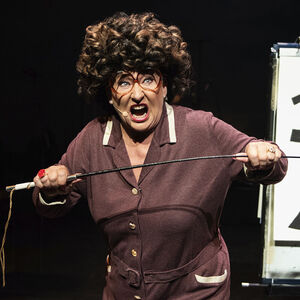 Karin Bloemen met een bruine pruik. Ze houdt een lange stok vast en kijkt boos de camera in. 