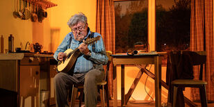 Een oudere man zit in een jaren 70 keuken en speelt banjo